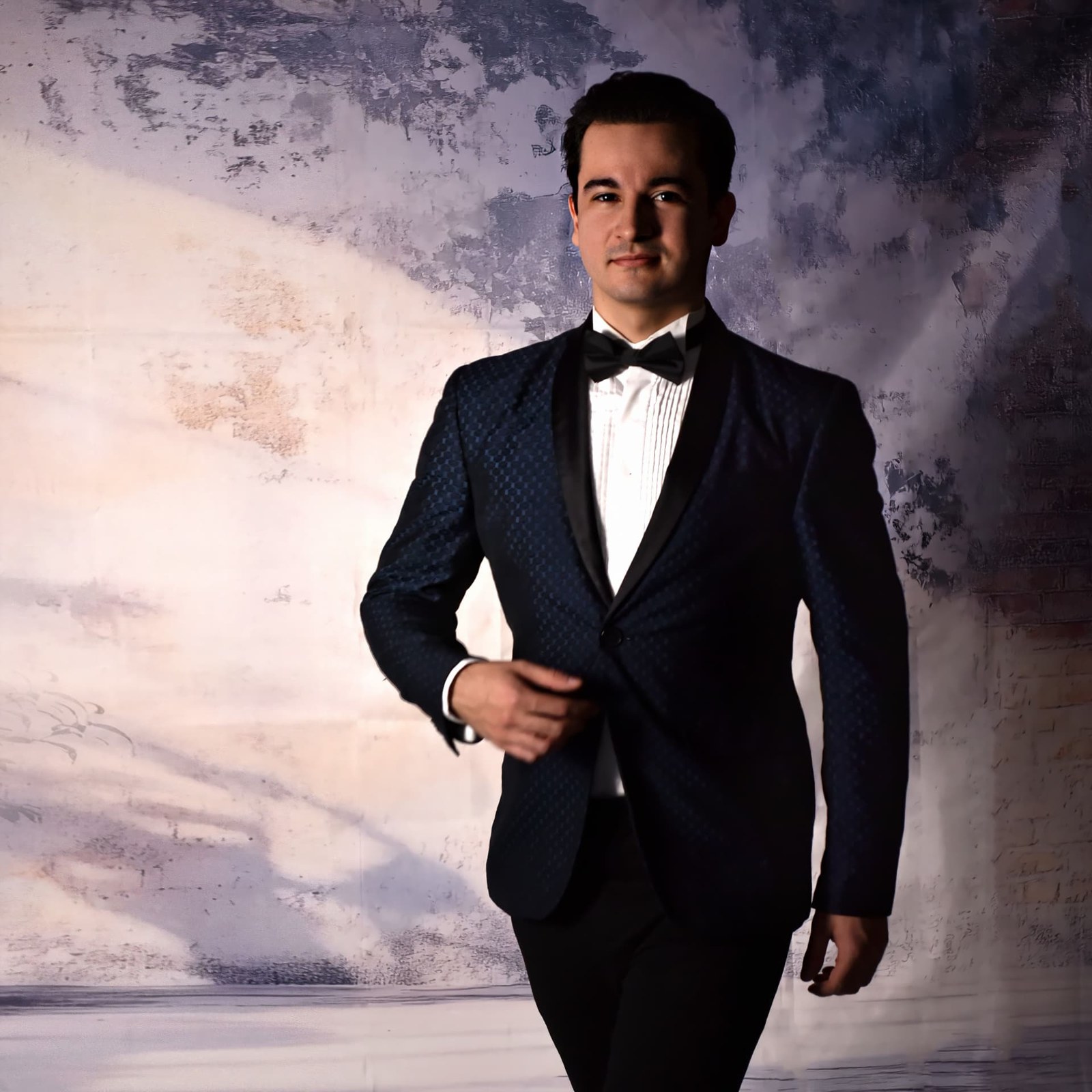 Jakub Osmera standing in a navy tuxedo against a painted wall